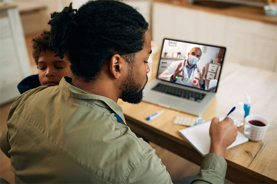 A patient takes notes from his telehealth care provider on a virtual visit; a child sleeps in his lap